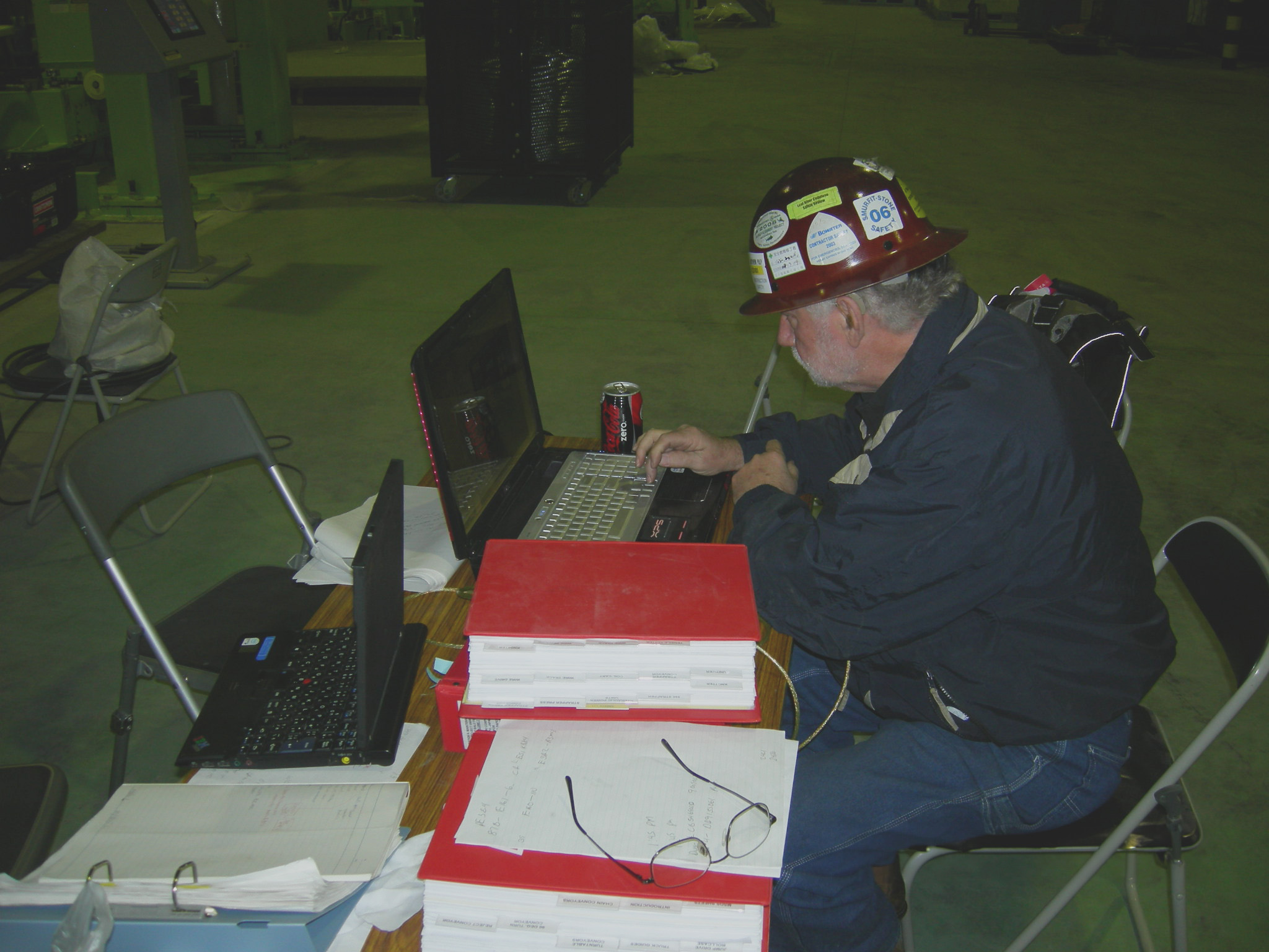 A man with a hard hat works on a laptop at a desk inside an industrial facility.