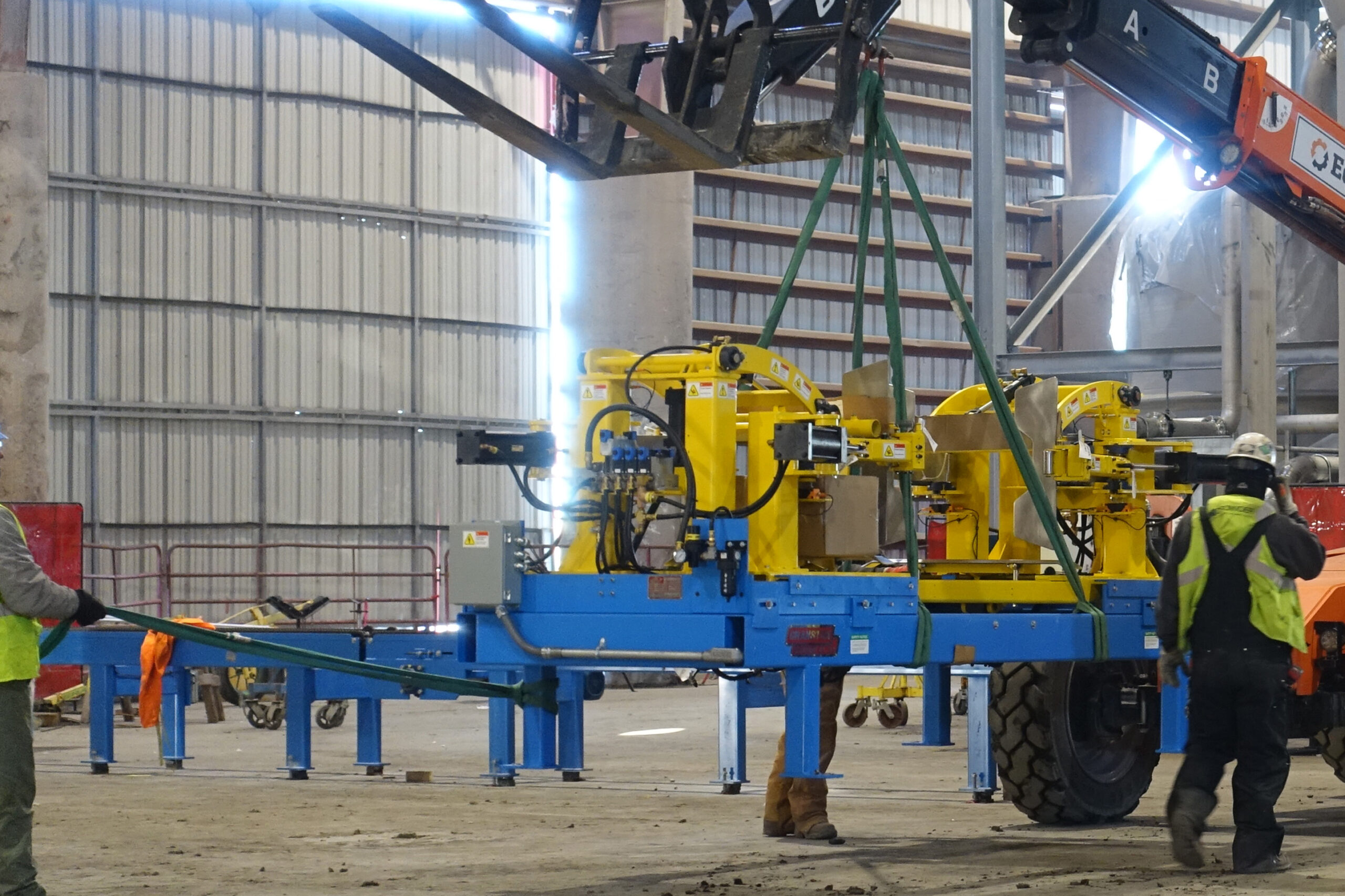 Workers guide a large yellow machine being lifted by a crane inside a warehouse.